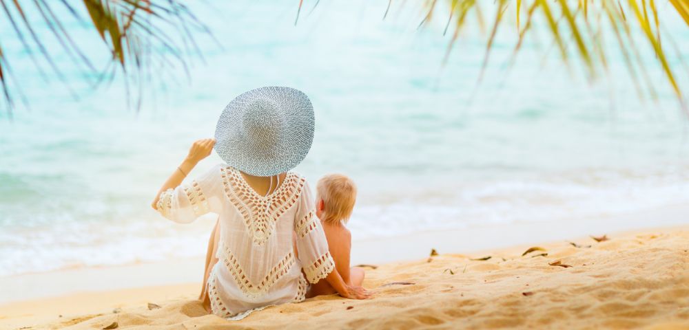 A person wearing a wide-brimmed hat and a white cover-up sits on a sandy beach next to a young child, both facing the ocean.