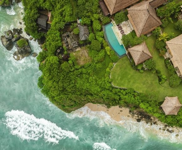 Aerial view of a tranquil tropical resort, with thatched-roof villas and a turquoise pool nestled in lush greenery beside a sandy, wave-lined shore.