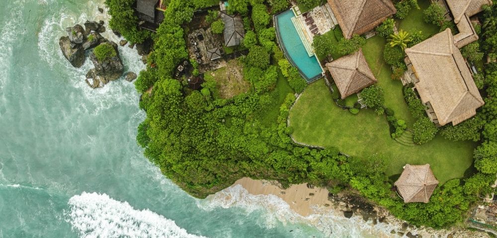 Aerial view of a tranquil tropical resort, with thatched-roof villas and a turquoise pool nestled in lush greenery beside a sandy, wave-lined shore.