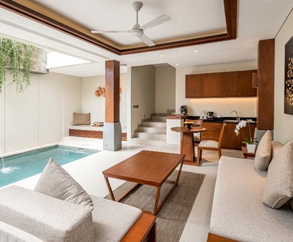 Bright modern open-plan living room with an indoor pool, pale sectional sofa, wood coffee table, compact kitchen, and tree artwork; calm and luxurious.