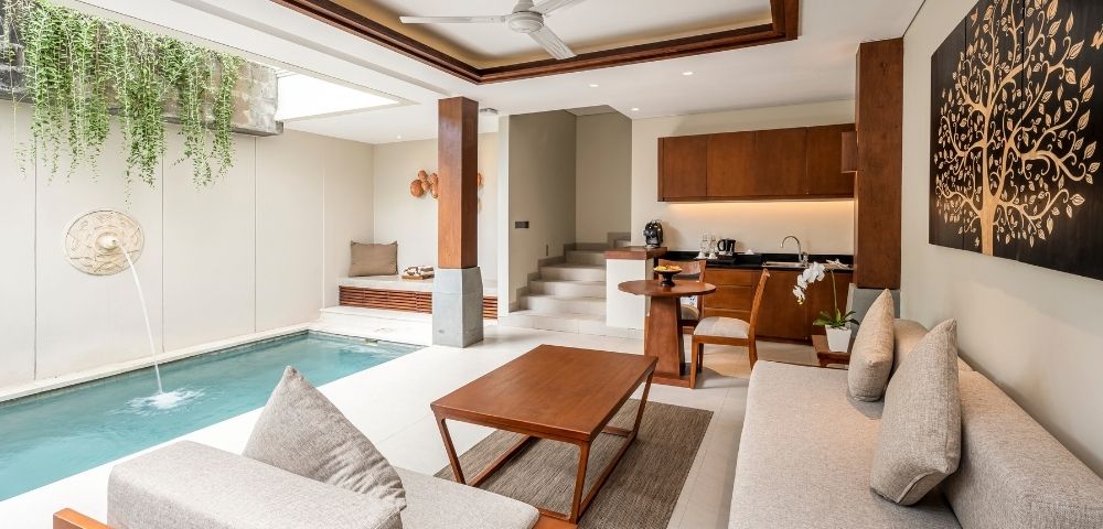 Bright modern open-plan living room with an indoor pool, pale sectional sofa, wood coffee table, compact kitchen, and tree artwork; calm and luxurious.