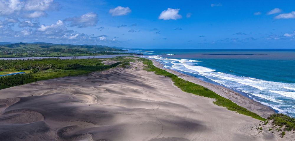 Aerial view of a vast, undulating sand dune system meeting a coastline with turquoise ocean waves crashing on the shore, framed by lush green vegetation and distant rolling hills under a bright blue sky with scattered clouds.