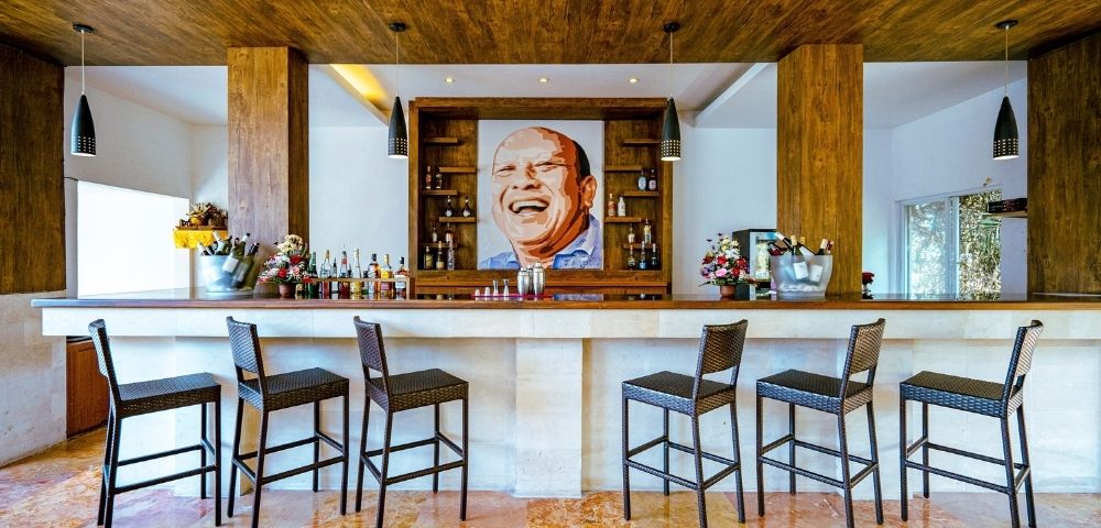 Interior view of a stylish bar area with a large portrait of a man laughing displayed prominently behind the bar. Several bar stools are neatly arranged in front of the bar counter, which is stocked with bottles and decorative items. The bar features warm wooden elements and ambient lighting.