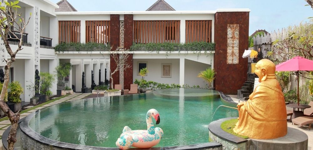 A tranquil resort courtyard with a large, green swimming pool, surrounded by modern architecture with stone and wood accents, and featuring a large golden Buddha statue with a fountain spout and a pink umbrella.