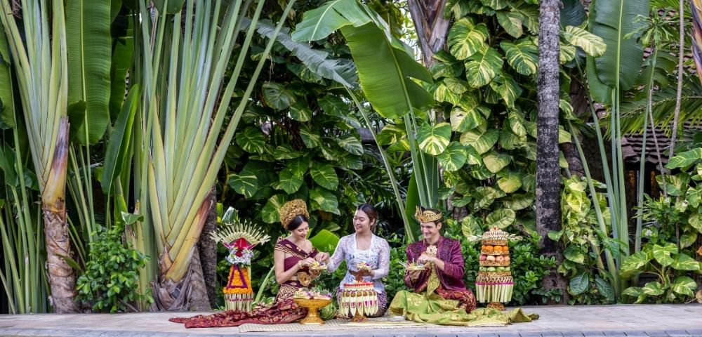 Three women in traditional dress sit on a stone platform amid lush tropical plants, with colorful fruit and flower offerings in a calm ceremonial scene.