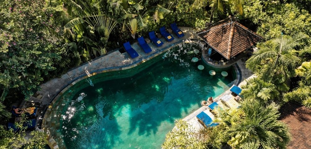 Aerial view of a turquoise resort pool surrounded by lush tropical palms, blue sun loungers, and a thatched-roof pavilion; serene, secluded atmosphere.