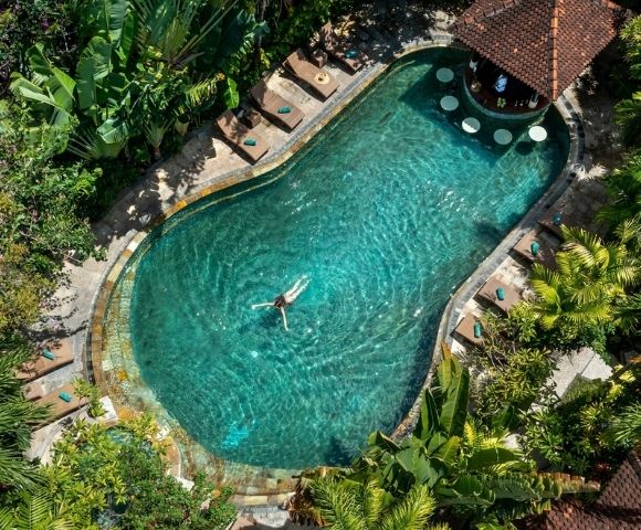 Aerial view of a turquoise resort pool surrounded by tropical greenery, with a lone swimmer, deck loungers, and a tiled-roof pavilion; serene and inviting.