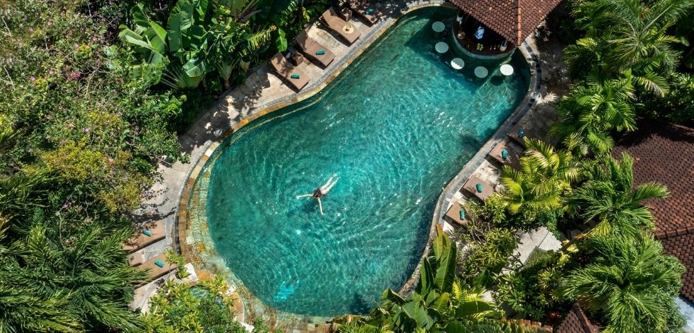Aerial view of a turquoise resort pool surrounded by tropical greenery, with a lone swimmer, deck loungers, and a tiled-roof pavilion; serene and inviting.
