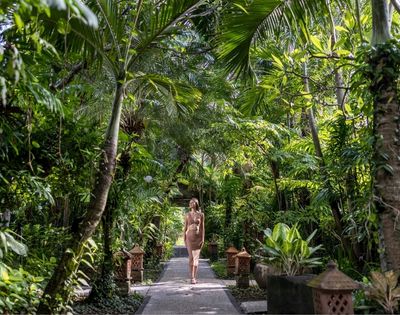 A woman walks along a stone path through a lush tropical garden, surrounded by palms, broad-leaf plants, and dense greenery; the scene feels serene and secluded.