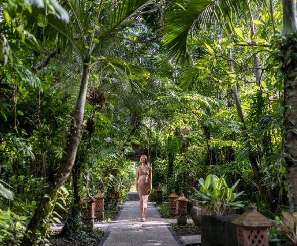 A woman walks along a stone path through a lush tropical garden, surrounded by palms, broad-leaf plants, and dense greenery; the scene feels serene and secluded.