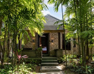 A secluded stone cottage with a tiled roof sits among lush tropical palms and garden foliage, its shadowed doorway creating a tranquil retreat.
