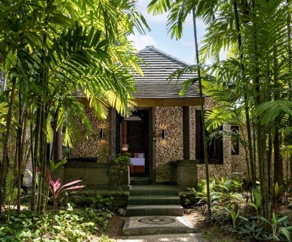 A secluded stone cottage with a tiled roof sits among lush tropical palms and garden foliage, its shadowed doorway creating a tranquil retreat.