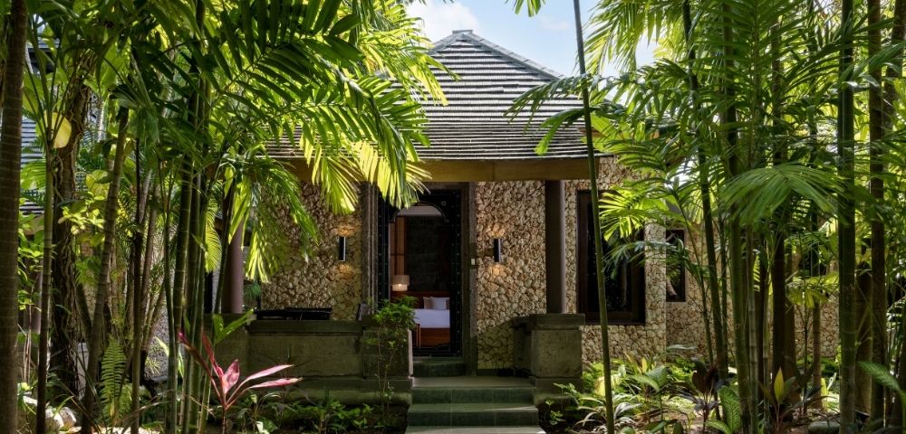 A secluded stone cottage with a tiled roof sits among lush tropical palms and garden foliage, its shadowed doorway creating a tranquil retreat.