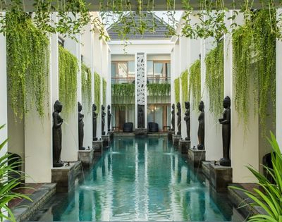 A tranquil courtyard pool reflects white columns and dark statues, framed by hanging vines and lush plants in an elegant, serene hotel setting.