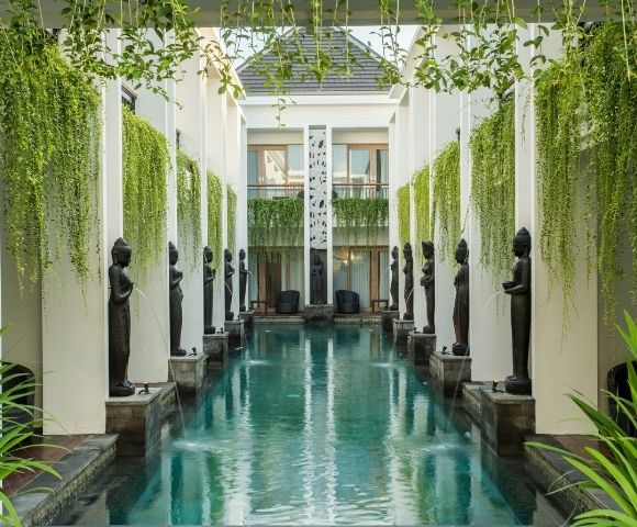 A tranquil courtyard pool reflects white columns and dark statues, framed by hanging vines and lush plants in an elegant, serene hotel setting.