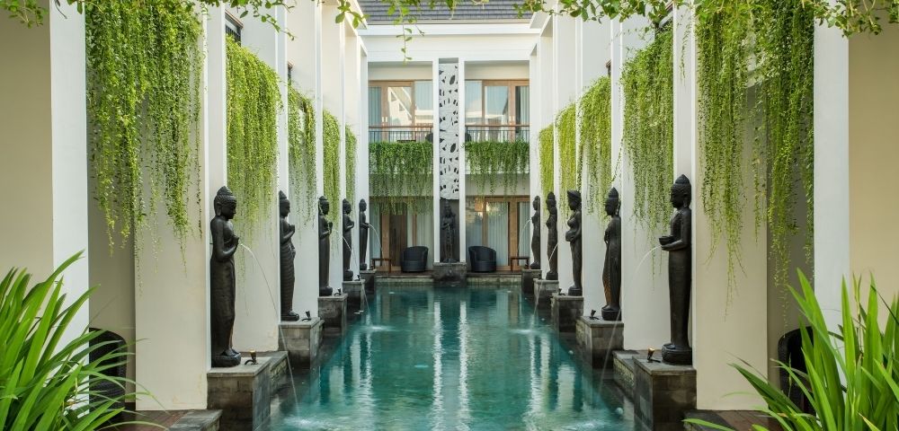 A tranquil courtyard pool reflects white columns and dark statues, framed by hanging vines and lush plants in an elegant, serene hotel setting.