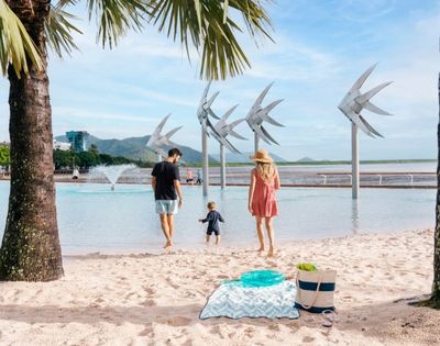 A family walks towards a man-made lagoon with large fish sculptures, with a palm tree in the foreground and a city skyline in the distance.