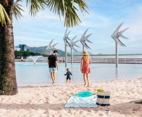 A family walks towards a man-made lagoon with large fish sculptures, with a palm tree in the foreground and a city skyline in the distance.