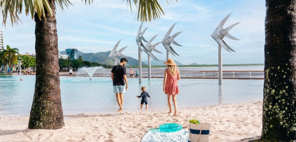 A family walks towards a man-made lagoon with large fish sculptures, with a palm tree in the foreground and a city skyline in the distance.