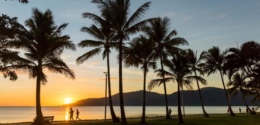 Silhouetted palm trees line a beach at sunset with two figures jogging along the water's edge.