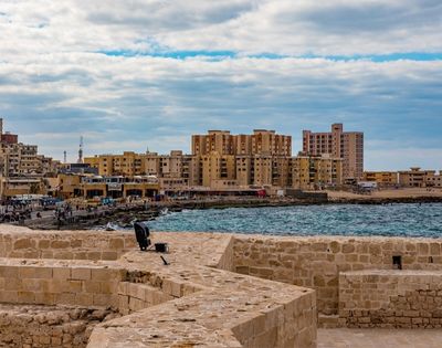 Ancient stone fortifications overlook a vibrant waterfront cityscape with modern buildings, a bustling promenade, and the shimmering blue sea under a cloudy sky.
