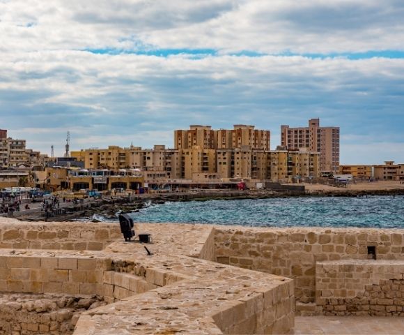Ancient stone fortifications overlook a vibrant waterfront cityscape with modern buildings, a bustling promenade, and the shimmering blue sea under a cloudy sky.