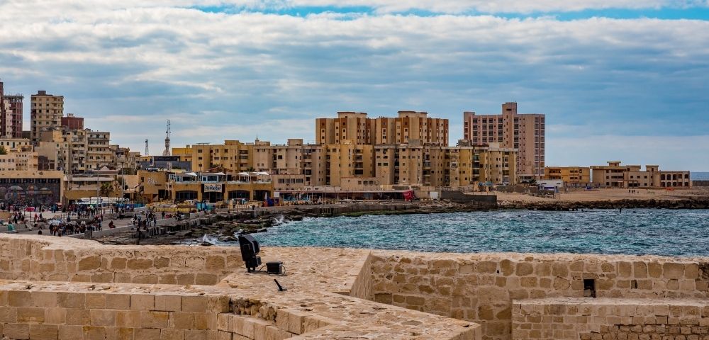 Ancient stone fortifications overlook a vibrant waterfront cityscape with modern buildings, a bustling promenade, and the shimmering blue sea under a cloudy sky.