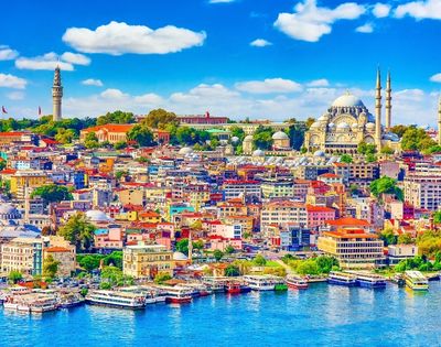 A vibrant, colorful cityscape of densely packed buildings with red roofs sits along a bright blue waterfront, with boats docked along the shore. In the background, a large domed mosque with minarets and a tall, slender tower dominate the skyline under a clear blue sky with fluffy white clouds.