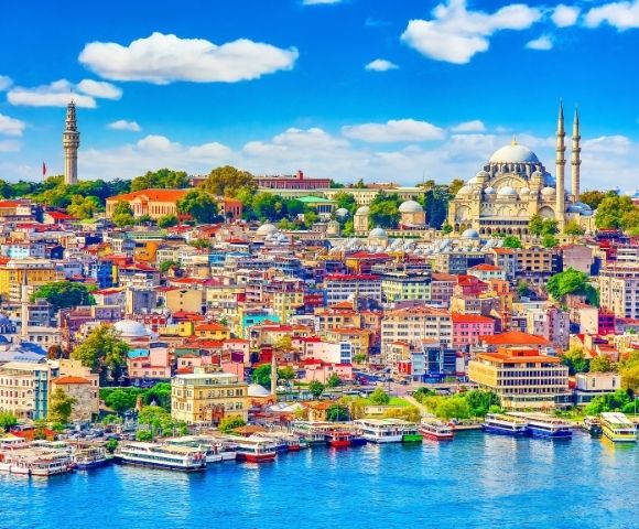 A vibrant, colorful cityscape of densely packed buildings with red roofs sits along a bright blue waterfront, with boats docked along the shore. In the background, a large domed mosque with minarets and a tall, slender tower dominate the skyline under a clear blue sky with fluffy white clouds.