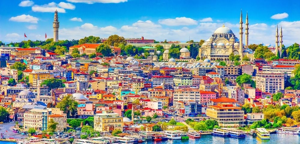 A vibrant, colorful cityscape of densely packed buildings with red roofs sits along a bright blue waterfront, with boats docked along the shore. In the background, a large domed mosque with minarets and a tall, slender tower dominate the skyline under a clear blue sky with fluffy white clouds.