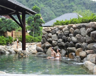 Two bathers relax in a clear outdoor hot spring beneath a timber pavilion, surrounded by a rugged stone wall and lush mountain greenery. The mood is peaceful and secluded.
