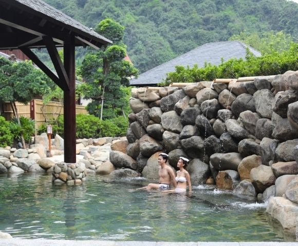 Two bathers relax in a clear outdoor hot spring beneath a timber pavilion, surrounded by a rugged stone wall and lush mountain greenery. The mood is peaceful and secluded.