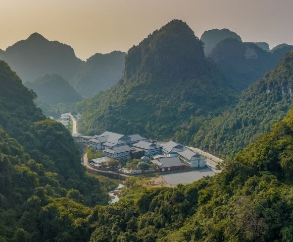 Aerial view of a small village nestled among lush, steep karst mountains, with clustered rooftops and a winding access road in soft, hazy light.
