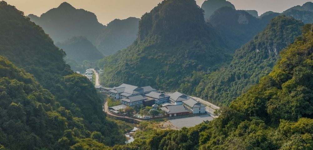 Aerial view of a small village nestled among lush, steep karst mountains, with clustered rooftops and a winding access road in soft, hazy light.