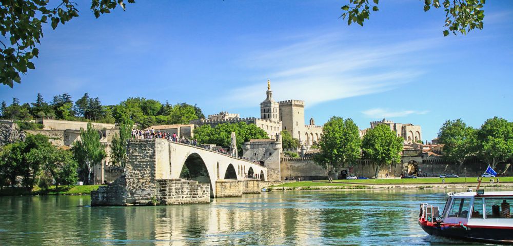 Stone bridge with arched spans crosses a wide river with a tour boat in the foreground and a historic palace complex on a hill in the background under a bright blue sky with wispy clouds.