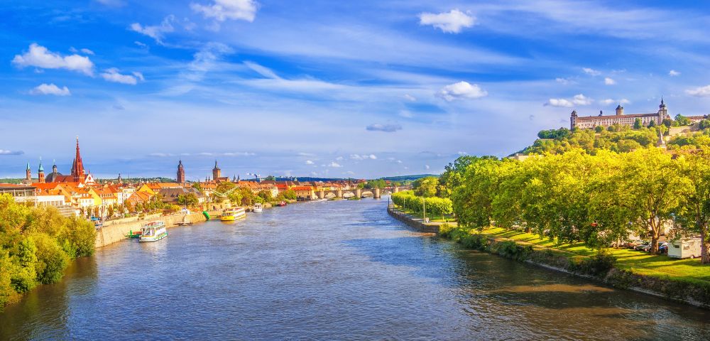 A wide, blue river flows through a city with historic architecture on the left bank and a tree-lined promenade leading to a large building on a hill to the right, under a bright blue sky with white clouds.