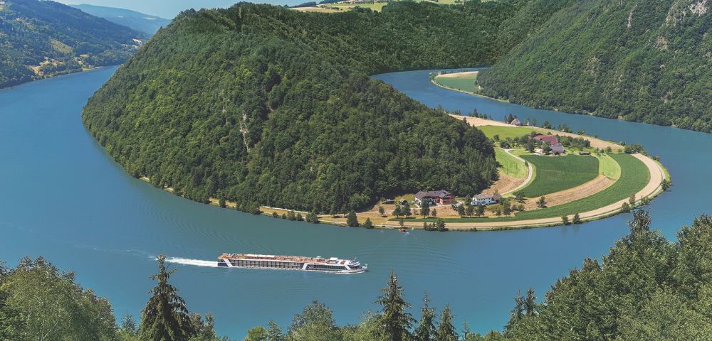 River cruise ship navigates a sharp bend in a wide, blue river surrounded by densely forested, rolling green hills and agricultural fields.