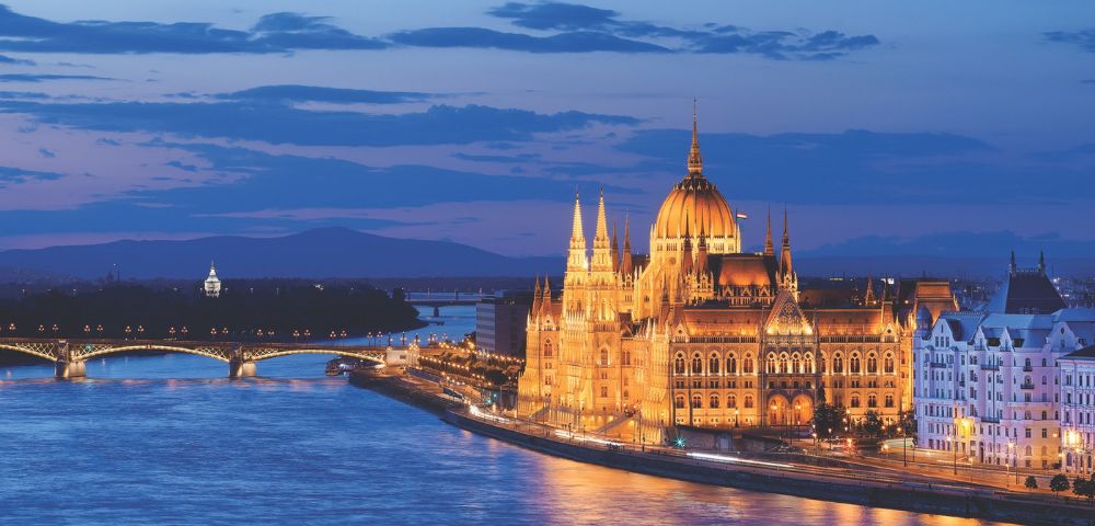 The illuminated Hungarian Parliament Building stands majestically along the Danube River at twilight, with a bridge and distant mountains visible in the background.