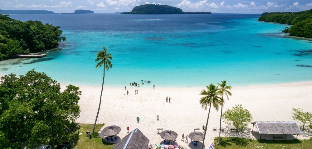 A tranquil tropical beach with turquoise water, white sand, palm trees, and distant green islands under a blue sky; visitors stroll along the shore.