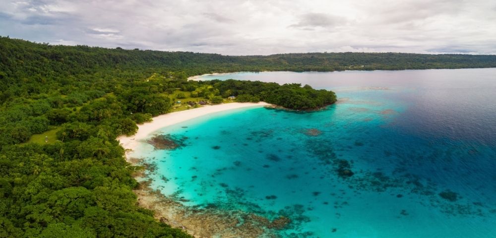 Aerial view of a quiet tropical bay, where brilliant turquoise water meets a crescent of white sand and dense green forest beneath a cloudy sky.
