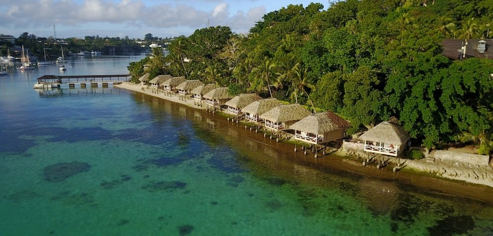 Aerial view of thatched bungalows along a lush tropical shore beside clear turquoise water, with boats in a quiet bay; a serene resort scene.