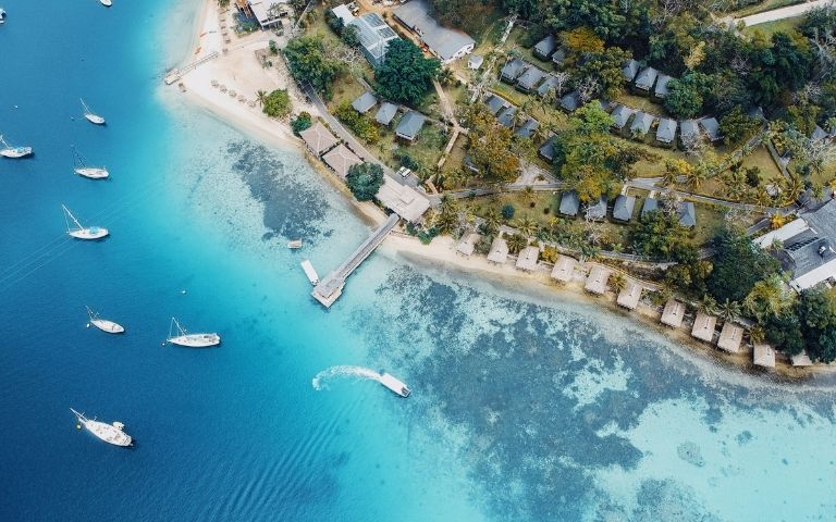 Aerial view of a peaceful tropical island, with turquoise water, anchored boats, a sandy beach, and small huts nestled among lush trees.