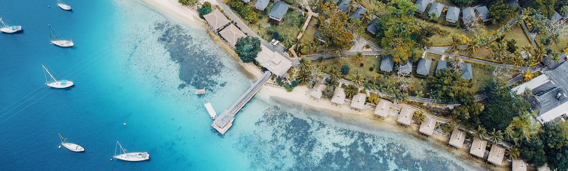Aerial view of a peaceful tropical island, with turquoise water, anchored boats, a sandy beach, and small huts nestled among lush trees.