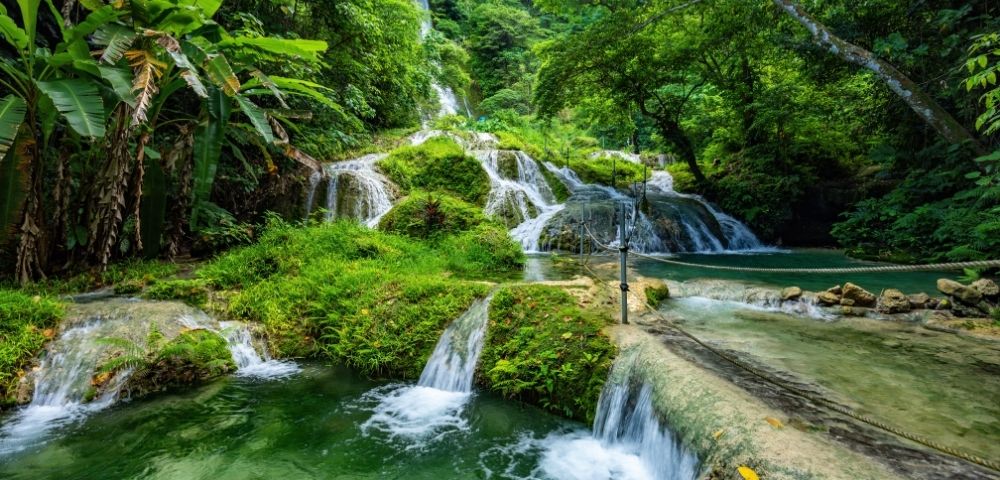 A tiered waterfall cascades through lush tropical rainforest into clear emerald pools, creating a vivid, tranquil scene surrounded by dense greenery.
