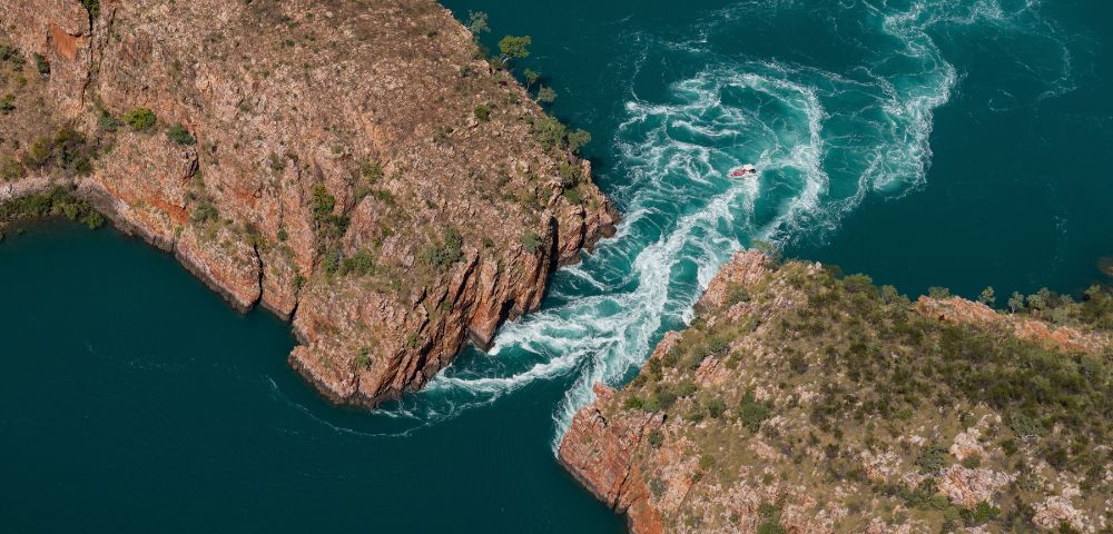 Aerial view of a small boat navigating turbulent turquoise waters flowing through a narrow gap between rugged, rocky cliffs dotted with sparse vegetation.
