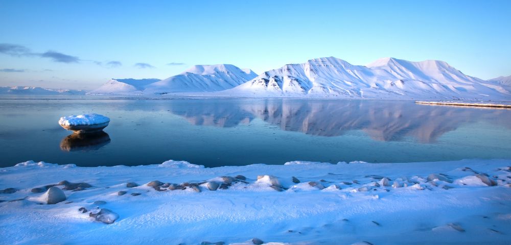 A serene Arctic landscape under a clear blue sky, featuring snow-capped mountains reflecting in calm, still water. A small, ice-covered rock sits in the foreground water.
