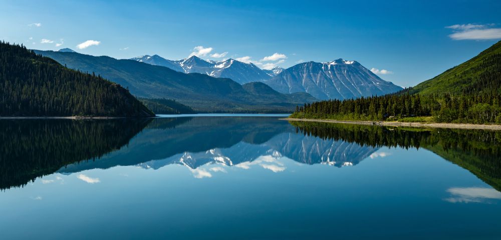A tranquil, mirror-like lake reflects snow-capped mountains and lush green forested shorelines under a clear blue sky.