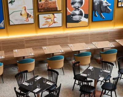 A warmly lit modern restaurant with wooden booths, small tables set with glasses and cutlery, teal-cushioned chairs, and colorful framed artwork.