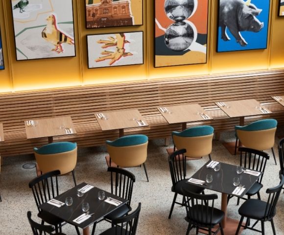 A warmly lit modern restaurant with wooden booths, small tables set with glasses and cutlery, teal-cushioned chairs, and colorful framed artwork.
