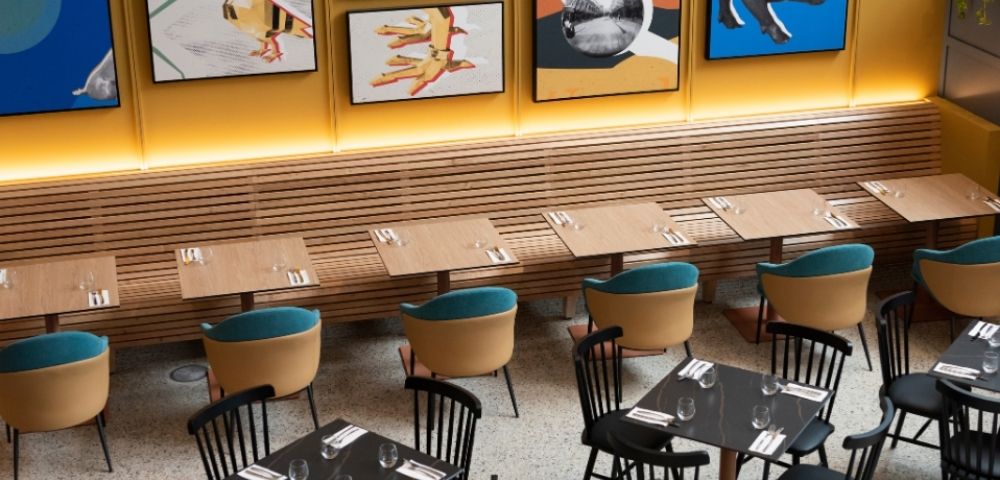 A warmly lit modern restaurant with wooden booths, small tables set with glasses and cutlery, teal-cushioned chairs, and colorful framed artwork.